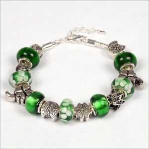 The Celtic Princess Bracelet, featuring a silver chain adorned with green and white glass beads along with various silver charms like a four-leaf clover, a butterfly, and a flower, ships quickly nationwide