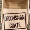 A Groomsman Crate - Wooden Gift Box with an open lid labeled "Groomsman Crate" in bold, black letters sits prominently on a granite countertop, appearing empty inside.